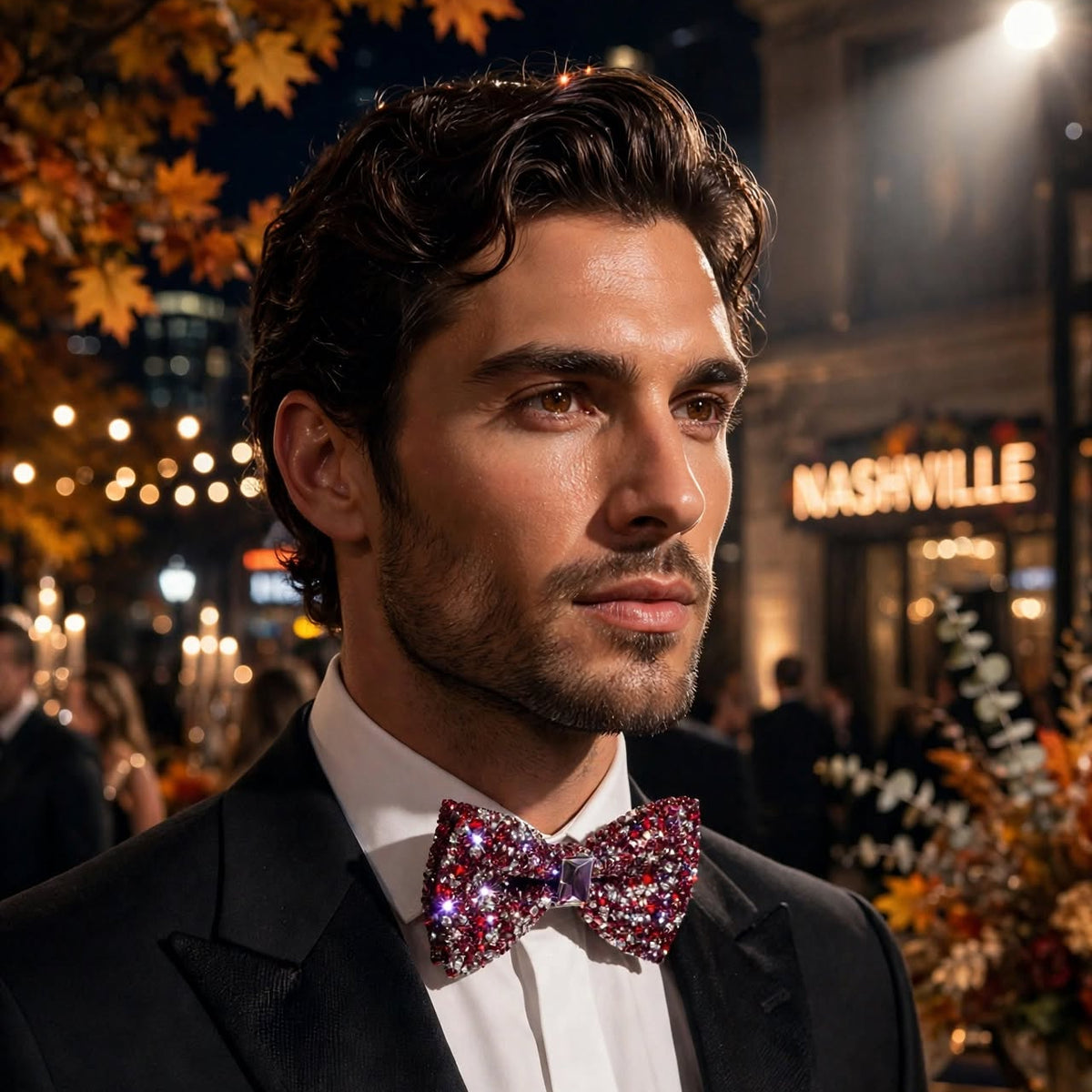 A man in a black tuxedo jacket and white dress shirt wearing a sparkling magenta and purple sequined bow tie, photographed outdoors at night with city lights and autumn foliage in the background.