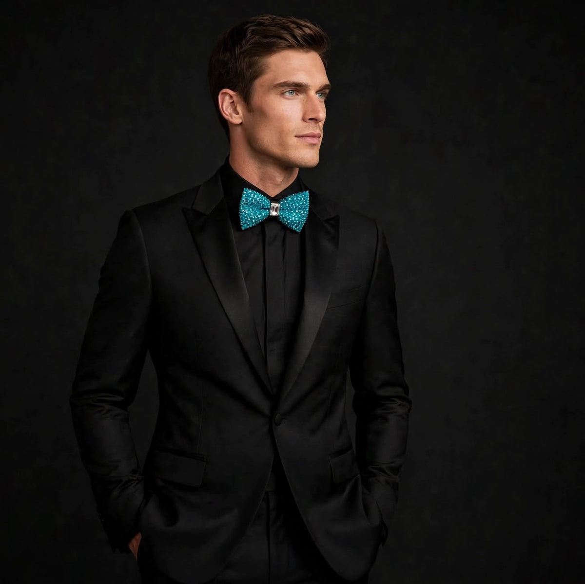 A man wearing a black tuxedo jacket with a black shirt and a bright turquoise patterned bow tie, posed against a dark textured studio background.